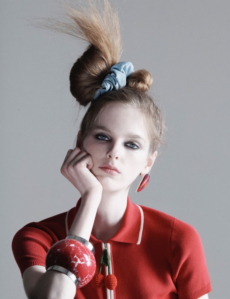 Portrait of a young adult female with an elaborate hairstyle, wearing a red collared shirt and large earrings, posing in a photo studio.