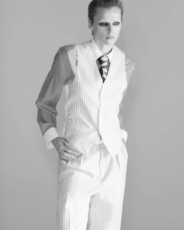 Fashion model in a pinstripe waistcoat and trousers with dark eye makeup posing against a grey studio backdrop.