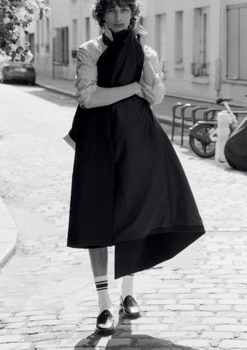 Model George Anderson wearing a striped shirt, draped black garment, white socks, and loafers on a cobblestone street in black and white.