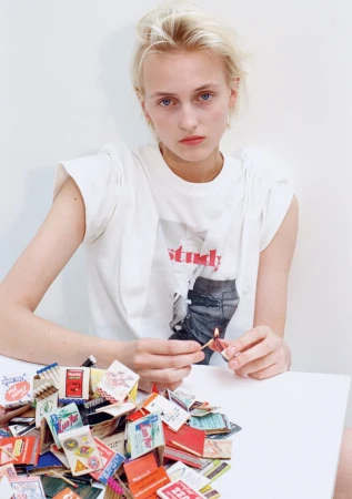 Model George Anderson in a white graphic t-shirt holding a lit matchstick above a pile of vintage matchbooks against a white studio wall.