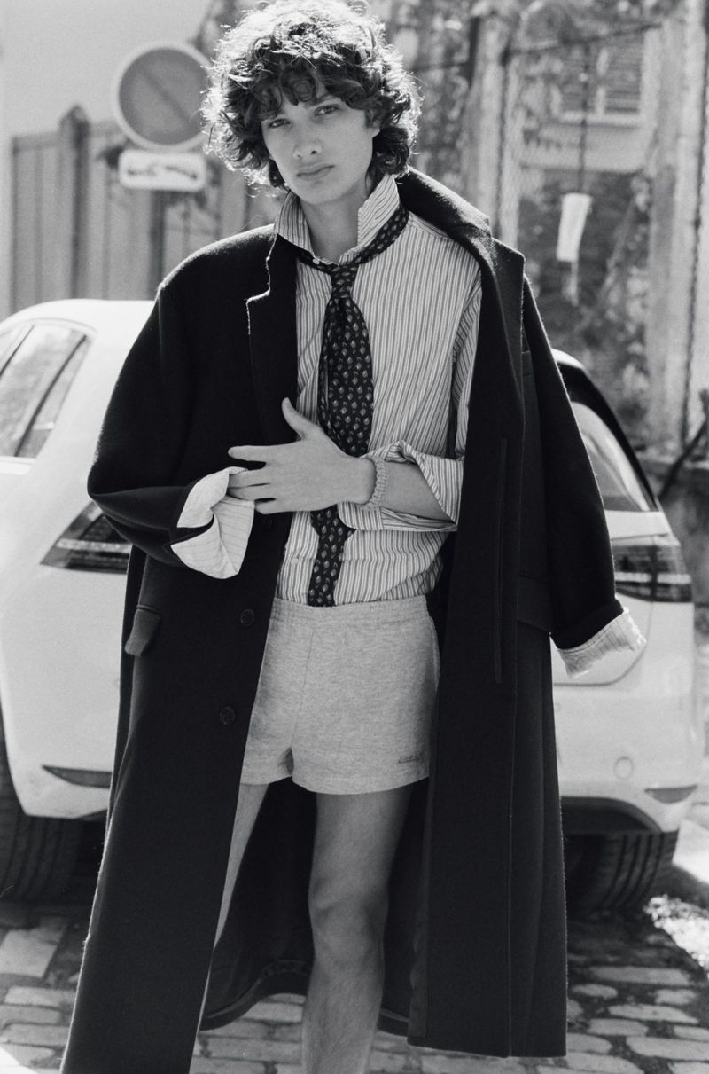 Model George Anderson wearing an oversized black coat, striped shirt, tie, and grey shorts on a cobblestone street.