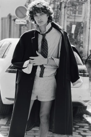 Model George Anderson wearing an oversized black coat, striped shirt, tie, and grey shorts on a cobblestone street.