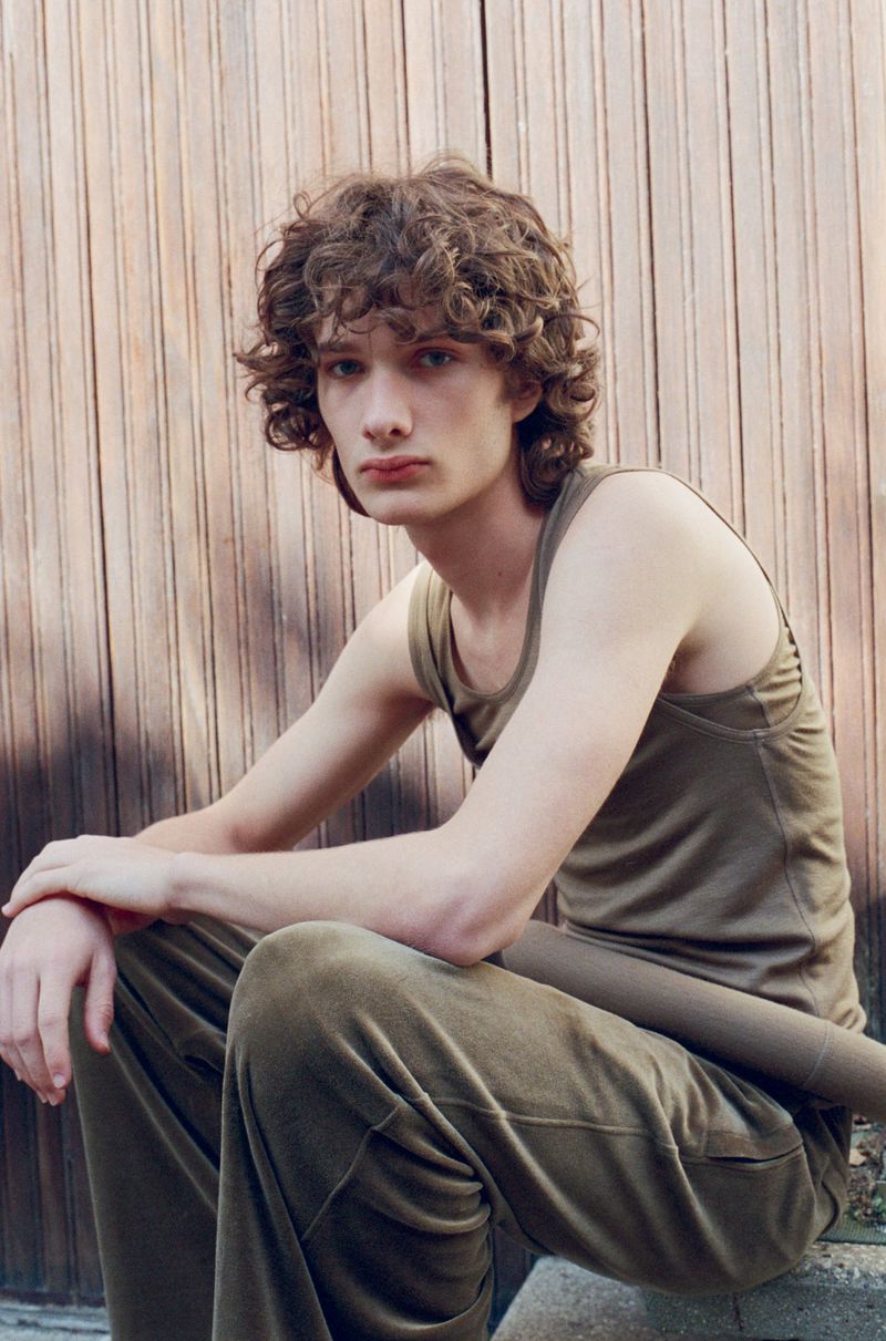 Model George Anderson seated in an olive green tank top and matching trousers in front of a vertical wood panel wall.