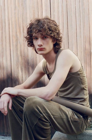 Model George Anderson seated in an olive green tank top and matching trousers in front of a vertical wood panel wall.