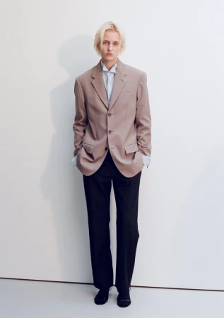 Model George Anderson wearing an oversized taupe blazer and black trousers against a neutral white studio backdrop.