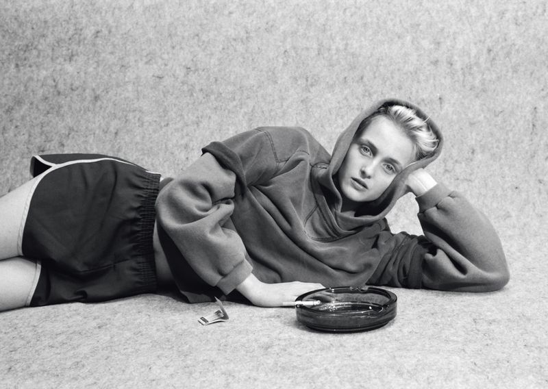 Model lying down on a textured surface wearing an oversized hoodie and athletic shorts next to an ashtray.