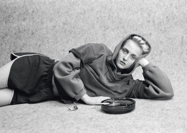 Model lying down on a textured surface wearing an oversized hoodie and athletic shorts next to an ashtray.