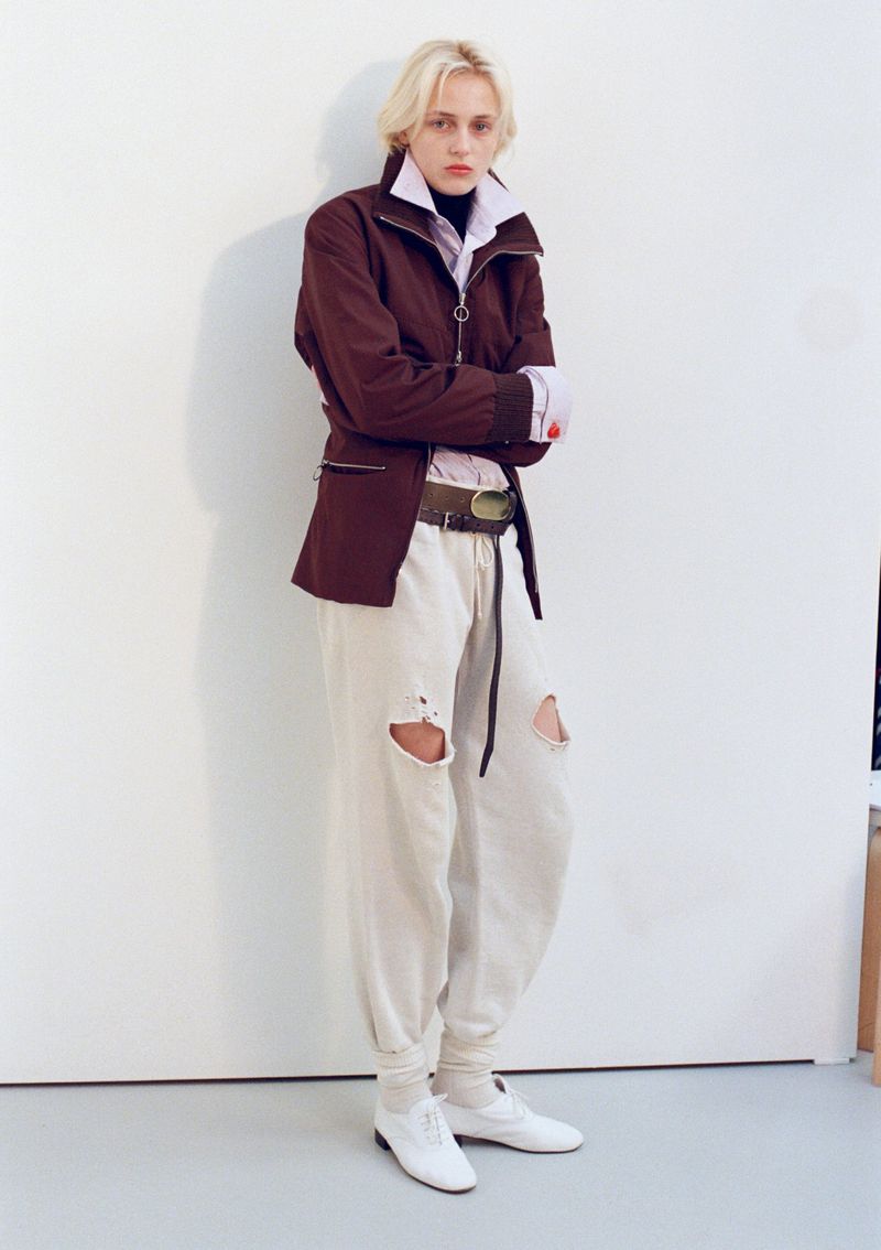 Model George Anderson posing against a white studio wall in a brown zip jacket, distressed sweatpants, and white shoes.