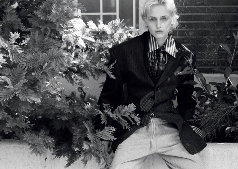 Model George Anderson wearing an oversized dark suit jacket, striped shirt, tie, and sweatpants outdoors among foliage.