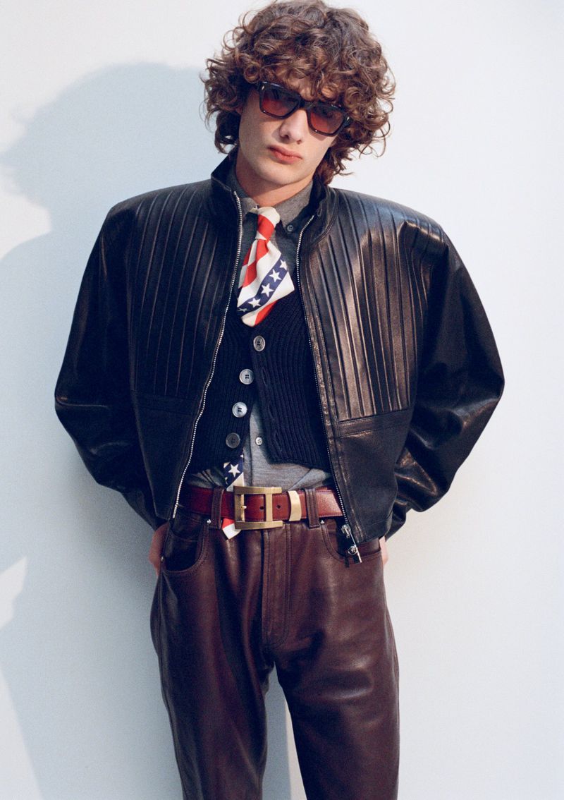 Model George Anderson wearing an oversized black leather jacket, patterned tie, and brown leather trousers against a white studio wall.