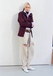 Model George Anderson posing against a white studio wall in a brown zip jacket, distressed sweatpants, and white shoes.