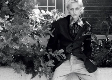 Model George Anderson wearing an oversized dark suit jacket, striped shirt, tie, and sweatpants outdoors among foliage.