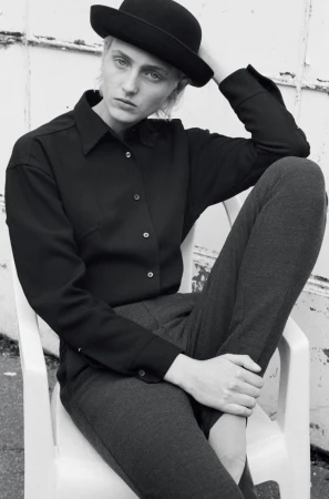 Model wearing a black button-down shirt, dark trousers, and a bowler hat seated in a white plastic chair outdoors.
