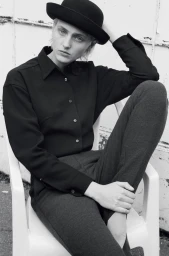 Model wearing a black button-down shirt, dark trousers, and a bowler hat seated in a white plastic chair outdoors.