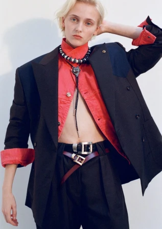 Model George Anderson wears an oversized black blazer over an open red shirt and layered belts against a white backdrop.