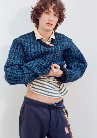 Model George Anderson lifts a blue patterned knit sweater over a striped collared shirt against a plain white studio backdrop.
