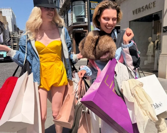 Two smiling women holding numerous designer shopping bags while walking along an outdoor retail street.
