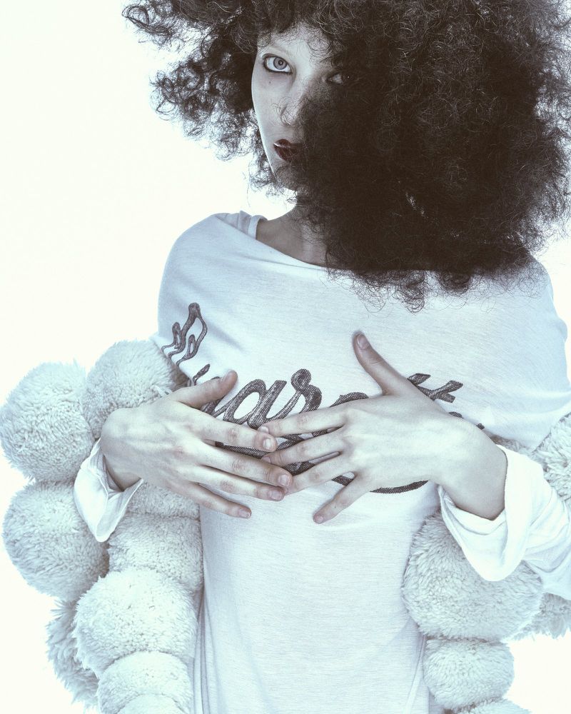 Model with voluminous dark curly hair wearing a white long-sleeve top wrapped in fluffy cream-colored circular pillows.