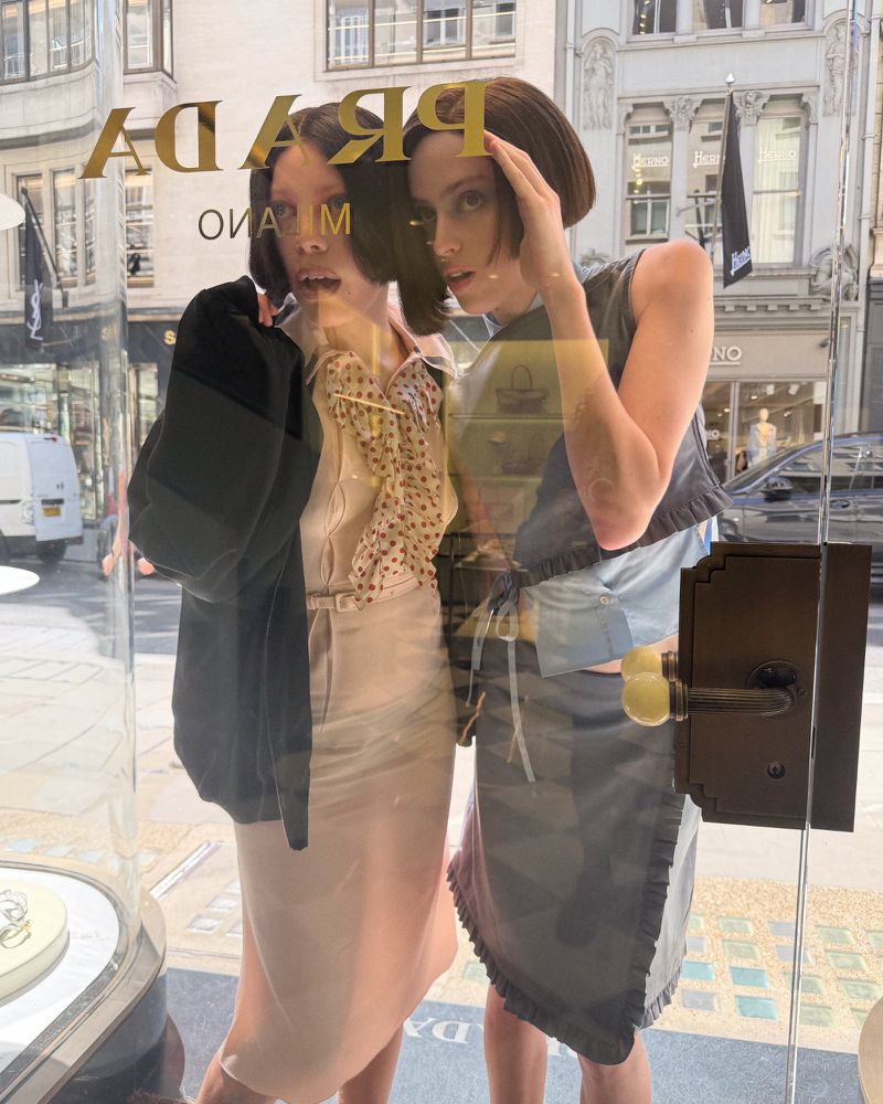 Two women with bob haircuts peer through a Prada glass window with street reflections across the surface.