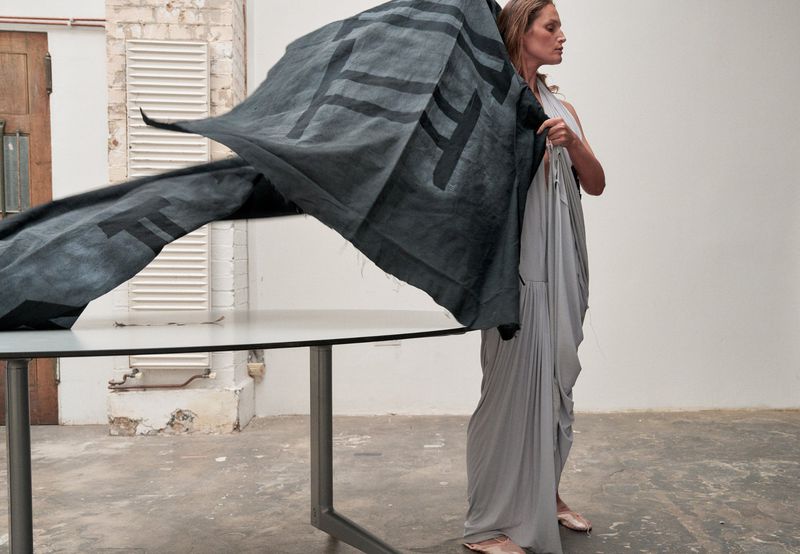 Vivien Solari wearing a draped light grey gown and holding a large graphic black fabric next to a table in a minimalist studio.