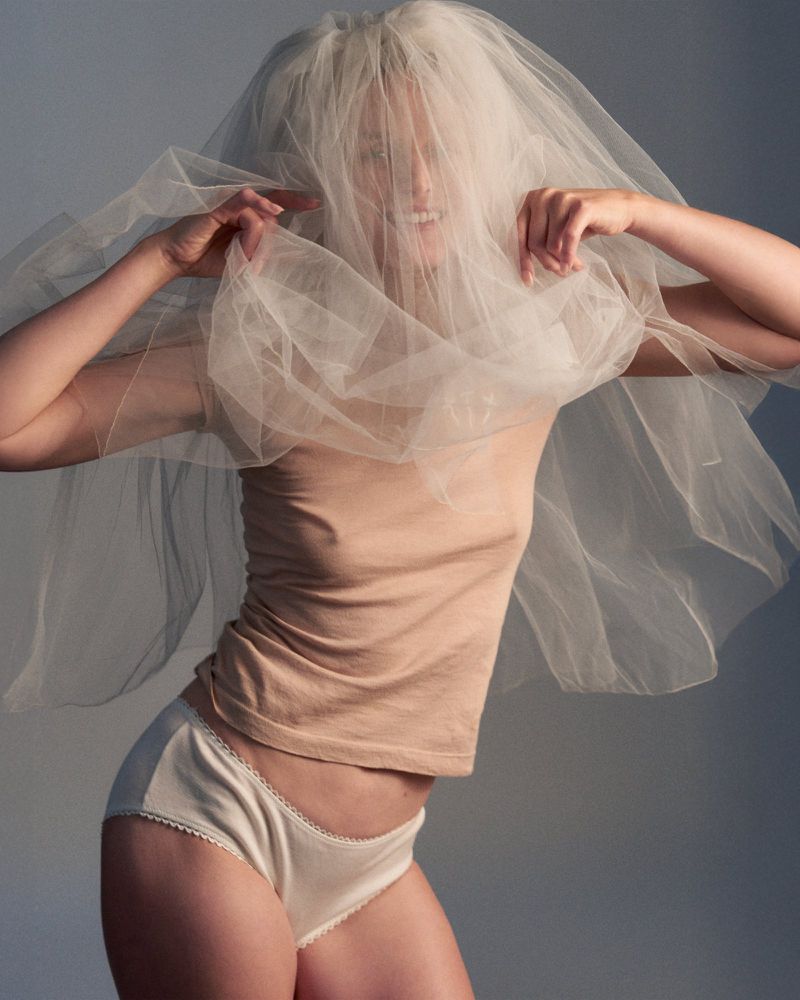 Model Vivien Solari covered in a voluminous sheer tulle veil, wearing a beige t-shirt and white briefs against a neutral studio backdrop.