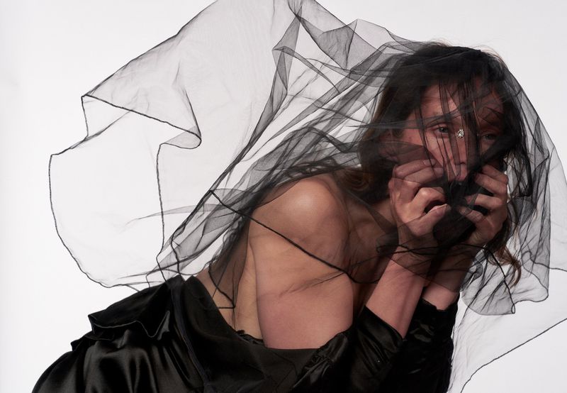 Model Vivien Solari draped in sheer black tulle netting over her face and wearing a black satin top against a white studio background.