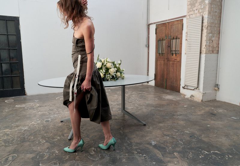 Model Vivien Solari wearing an olive strapless midi dress and mint green pumps next to an oval table with a white flower bouquet.