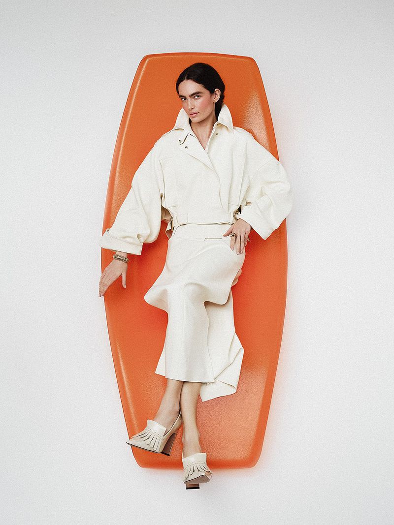 Model wearing white jacket and skirt reclining on a bright orange surfboard against a seamless white background.