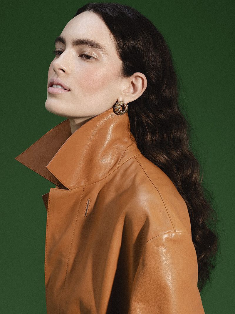Woman in a high-collared caramel leather coat and hoop earring posing in profile against a dark green backdrop.