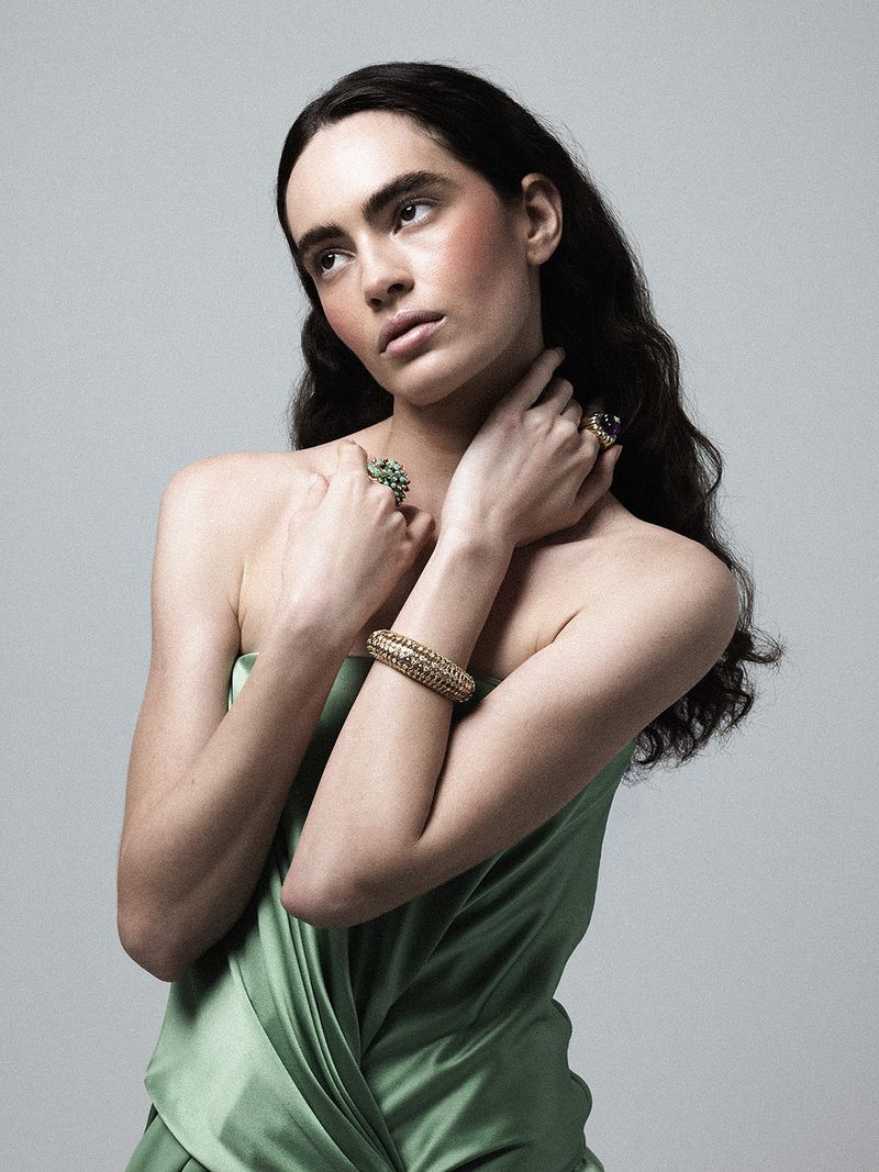 Model wearing a strapless light green satin dress, textured gold bracelet, and gemstone rings against a grey studio backdrop.