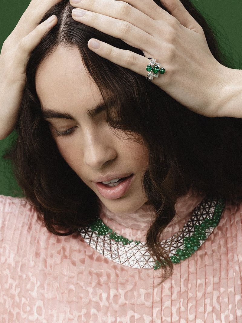 Close-up portrait of a woman wearing an emerald and diamond necklace and ring with hands in her dark hair against a green backdrop.