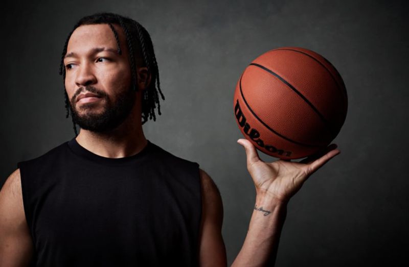 Jalen Brunson in a black sleeveless shirt balancing an orange basketball in his hand against a dark grey textured backdrop.