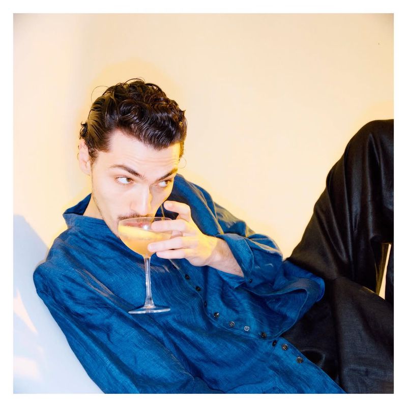 Man wearing a blue button-down shirt holding a coupe glass against a plain light wall.