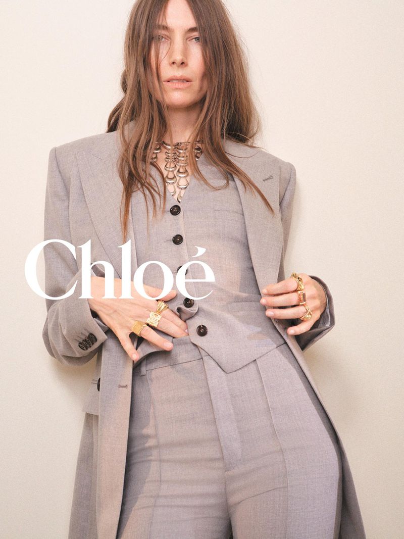Model Jessica Miller wearing a tailored grey three-piece suit and bold gold jewellery for Chloé against an off-white backdrop.