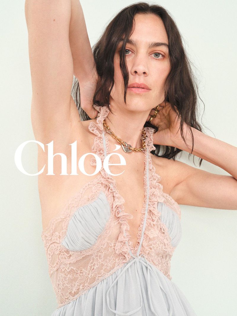Alexa Chung posing with hands behind her head in a ruffled pink lace halter dress against a pale studio backdrop.