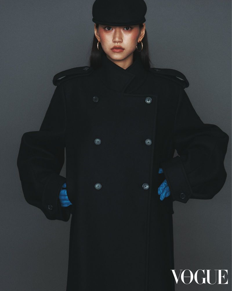 Woman in black coat and cap posing in a studio with gray background. The Vogue logo is visible in the bottom right corner.