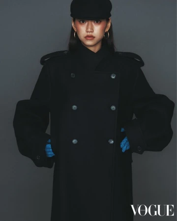 Woman in black coat and cap posing in a studio with gray background. The Vogue logo is visible in the bottom right corner.