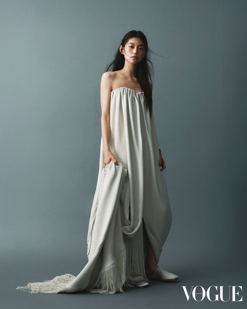 An Asian model poses in a strapless white dress with a fringed white throw in a studio setting against a blue background.