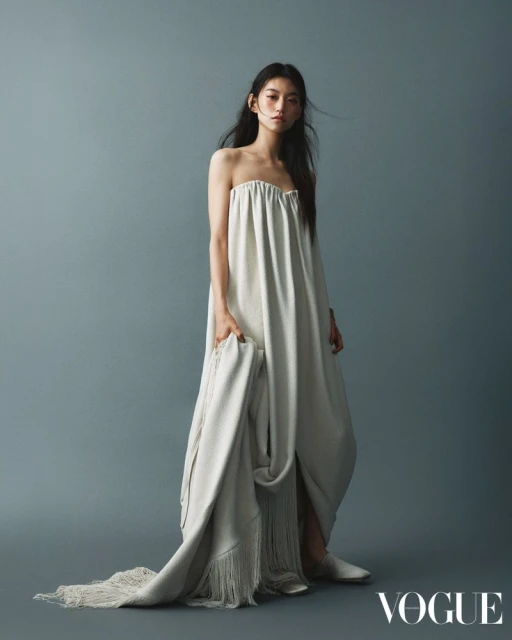 An Asian model poses in a strapless white dress with a fringed white throw in a studio setting against a blue background.