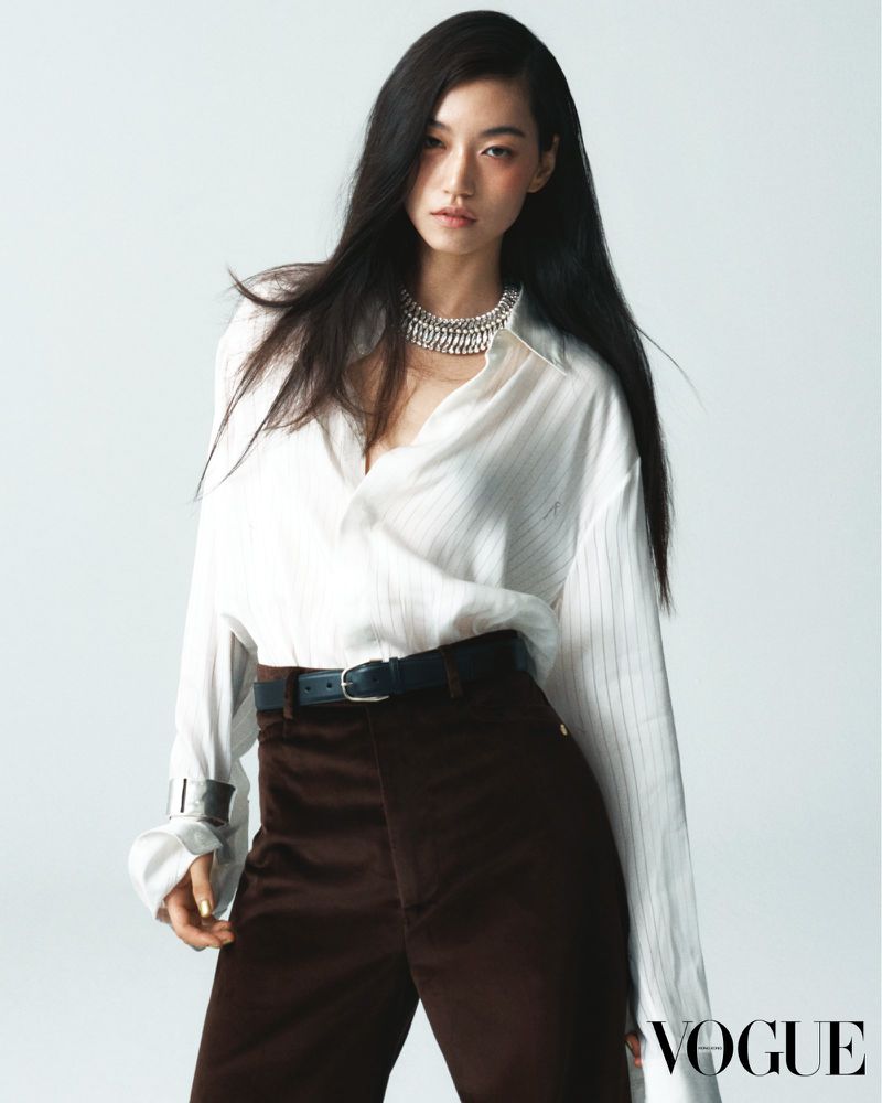 Fashion editorial shot of an Asian woman posing in a white striped shirt, brown pants, and a silver necklace against a white backdrop.