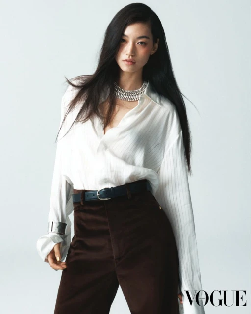 Fashion editorial shot of an Asian woman posing in a white striped shirt, brown pants, and a silver necklace against a white backdrop.