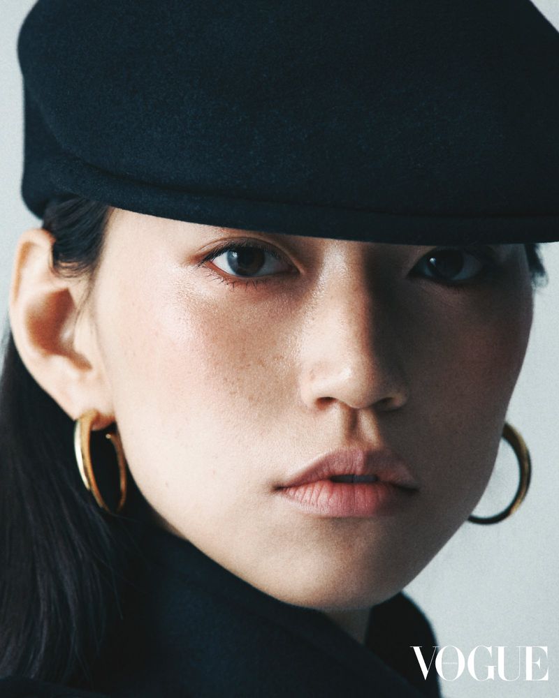Close-up studio portrait of an Asian adult female model wearing a black hat, gold hoop earrings, and black top. Vogue logo is visible.