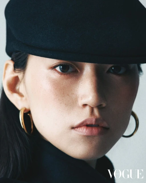 Close-up studio portrait of an Asian adult female model wearing a black hat, gold hoop earrings, and black top. Vogue logo is visible.