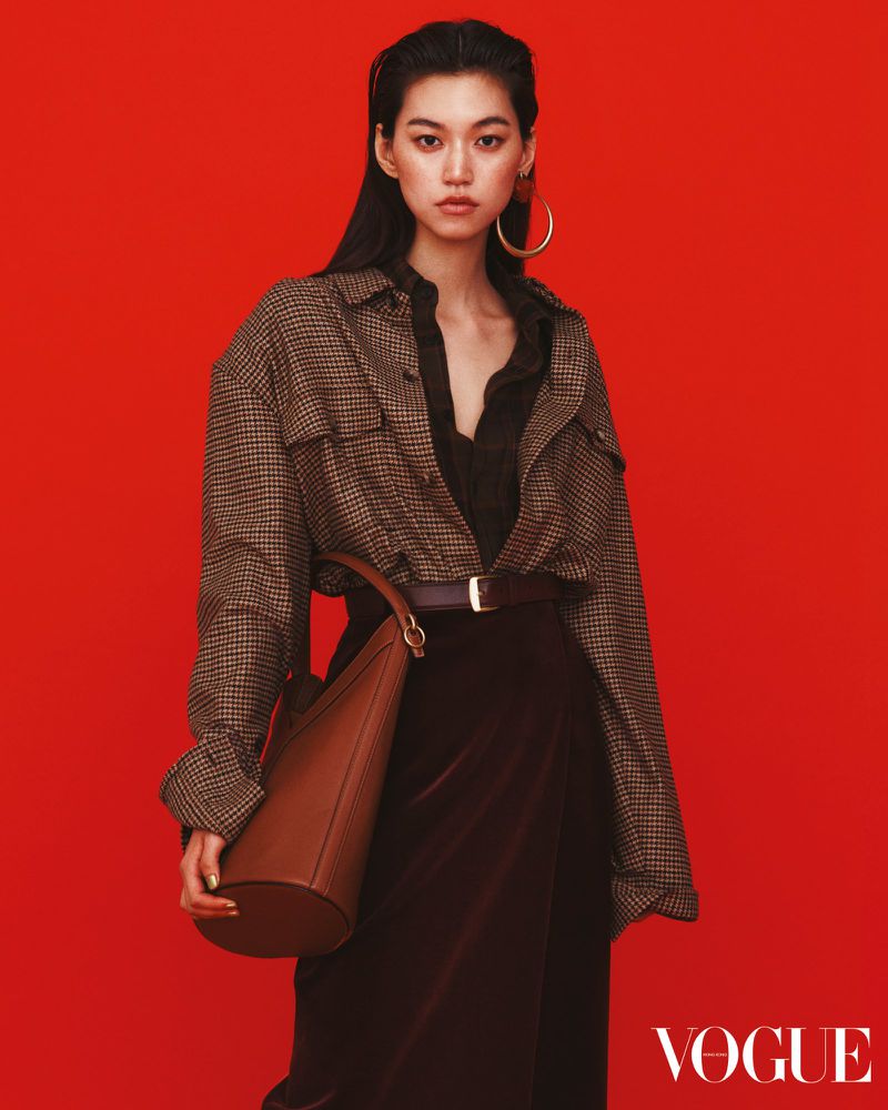 Asian female model in an oversized houndstooth jacket, brown skirt, and hoop earrings poses against a vibrant red backdrop.