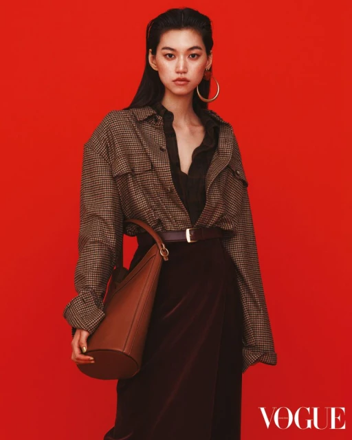 Asian female model in an oversized houndstooth jacket, brown skirt, and hoop earrings poses against a vibrant red backdrop.