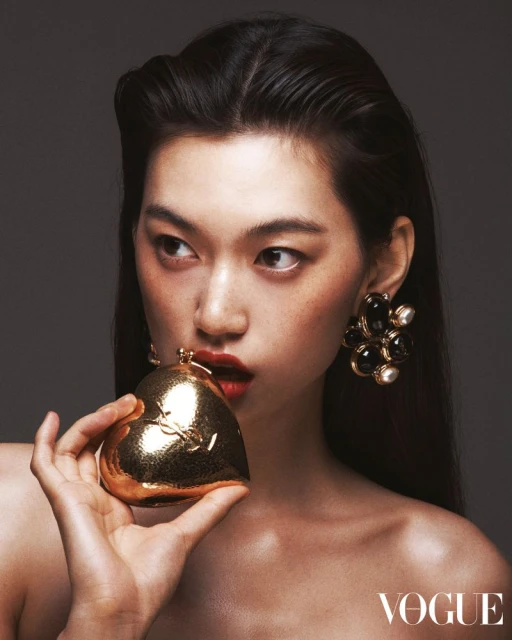 Glamour shot of a stylish Asian woman with flawless skin and dark hair, holding a gold heart-shaped purse and wearing large earrings.