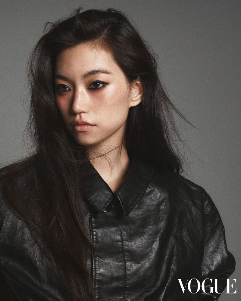 Close-up studio shot of an Asian woman wearing a black leather jacket against a grey backdrop with the Vogue logo in the corner.