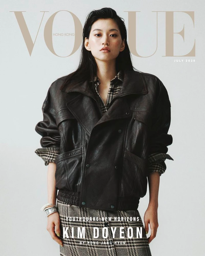 Kim Doyeon posing on the Vogue Hong Kong magazine cover wearing an oversized black leather jacket and plaid skirt against a pale backdrop.