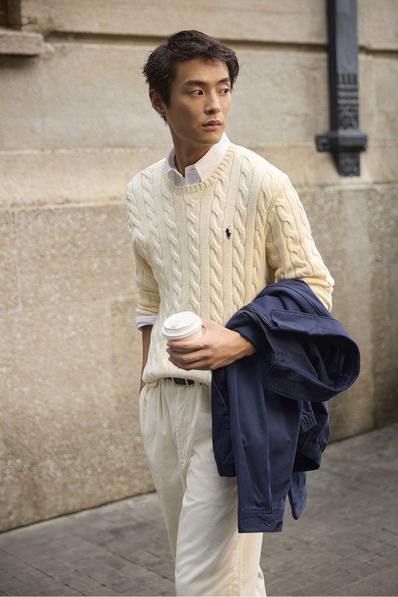 An East Asian man in a cream cable-knit sweater and white trousers holding a coffee cup and carrying a navy jacket outdoors.