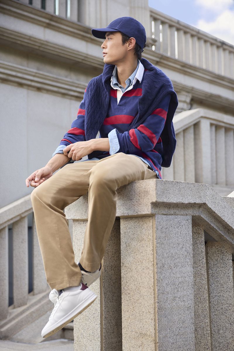 Man in a striped navy and red rugby shirt, beige chinos, blue cap, and white sneakers sitting on a stone balustrade.